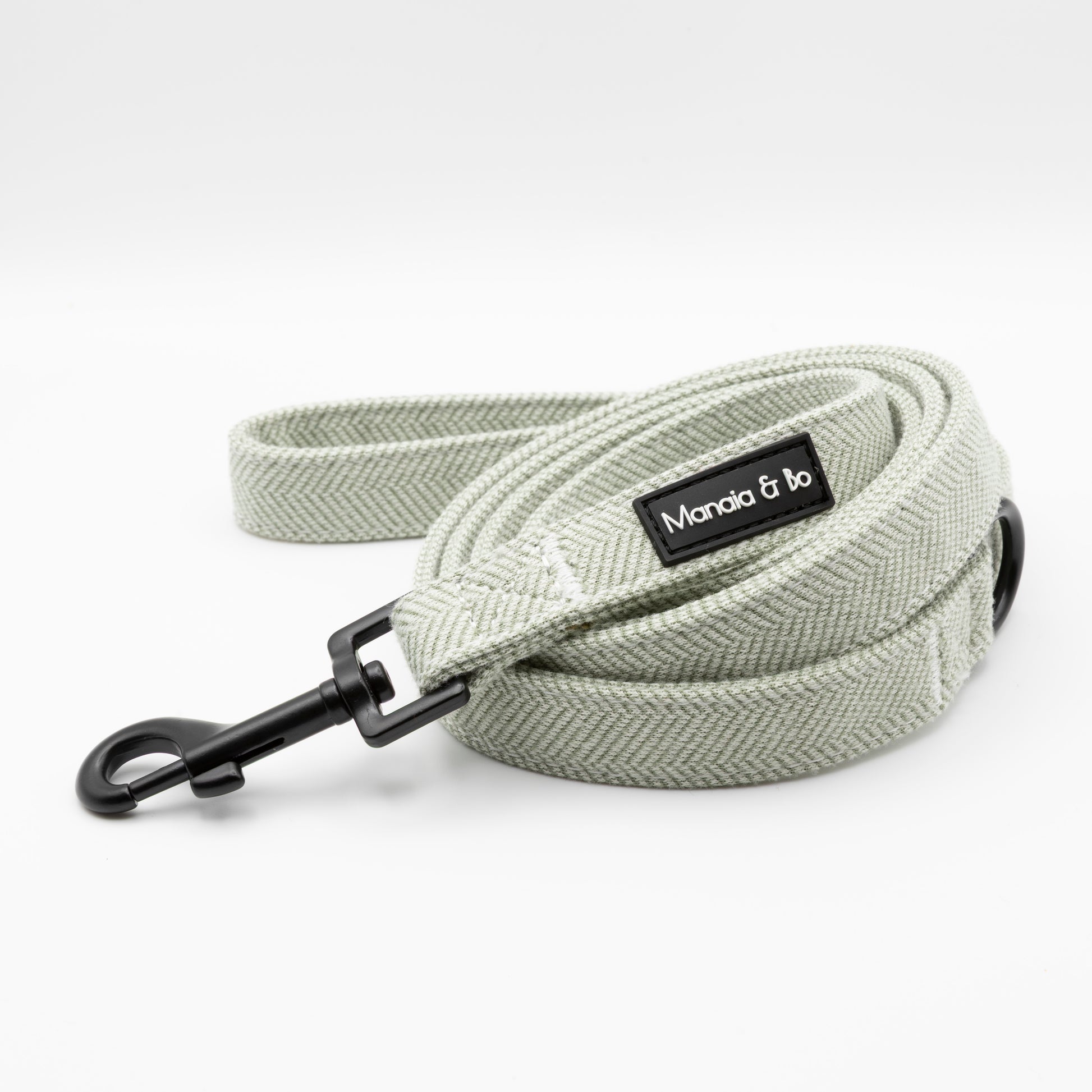 The Pine Tweed Harness & Lead features a light gray woven dog leash with a black metal clasp and a Manola & Bo label, coiled neatly on a white background—pair it with our adjustable harness for superior style and comfort.