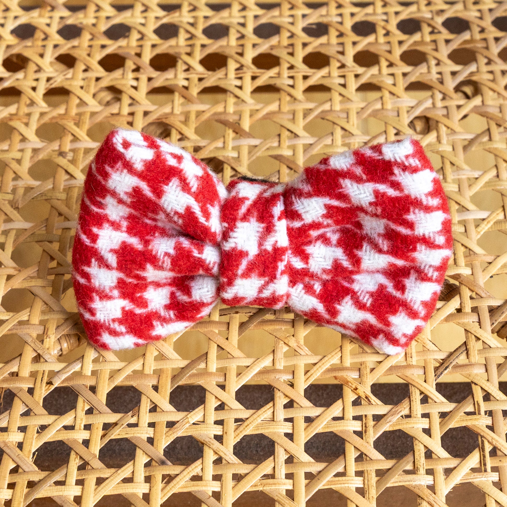 The Scarlet Houndstooth Dog Bow Tie in red and white lies on a woven rattan surface with a crisscross pattern.