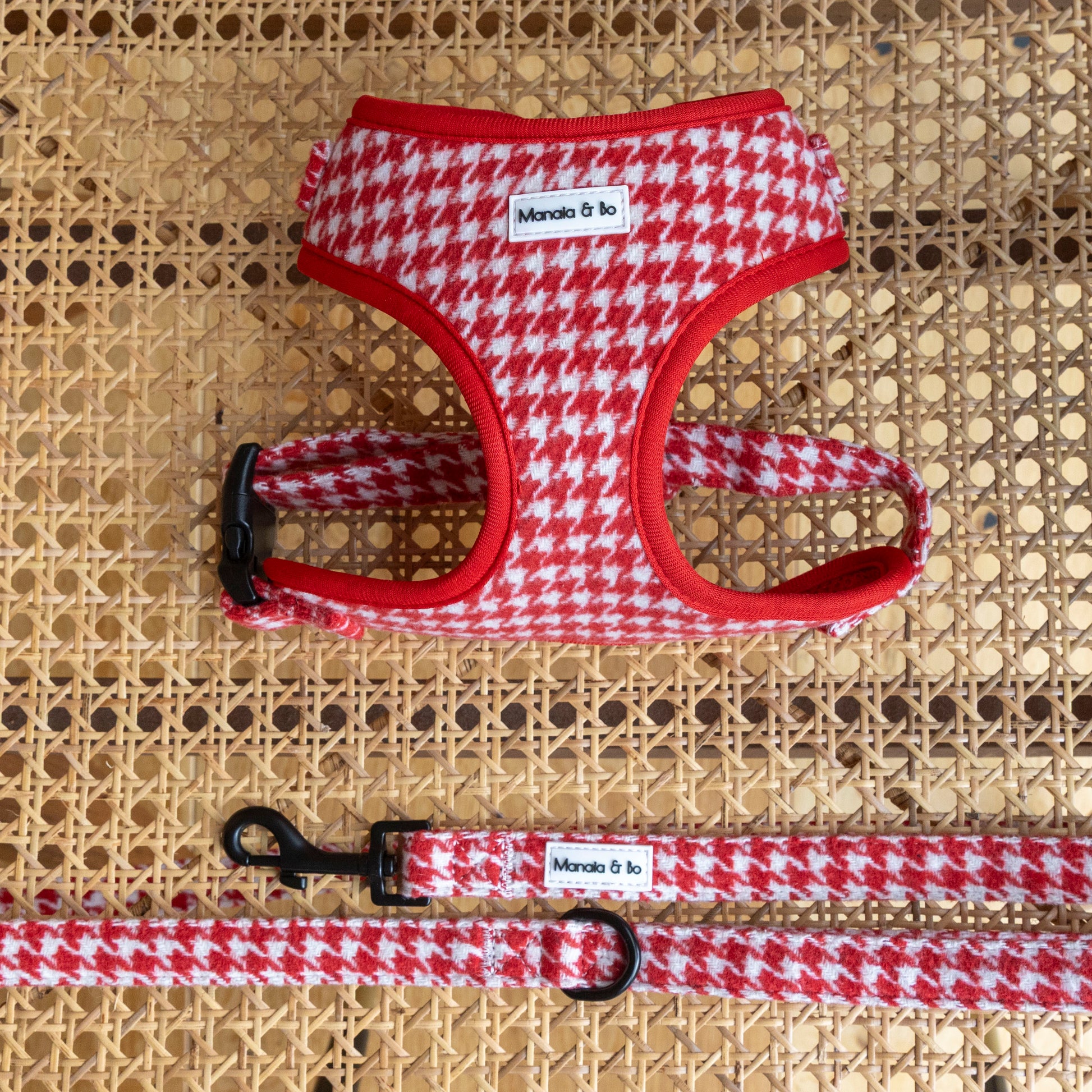 The Scarlet Houndstooth Harness & Lead set, featuring red and white houndstooth with black clips and Manada & Co. labels, rests on woven rattan. This adjustable harness ensures both style and comfort for your dog.