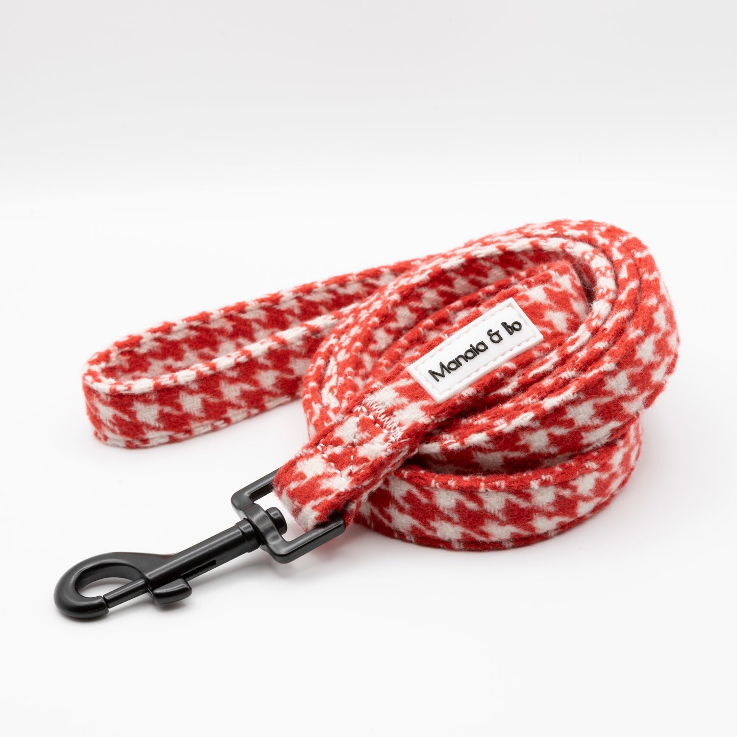 The Scarlet Houndstooth Dog Lead, featuring a red and white houndstooth pattern, black metal clasp, and a sewn-on fabric label with text, is coiled on a white background—ideal for stylish pups.