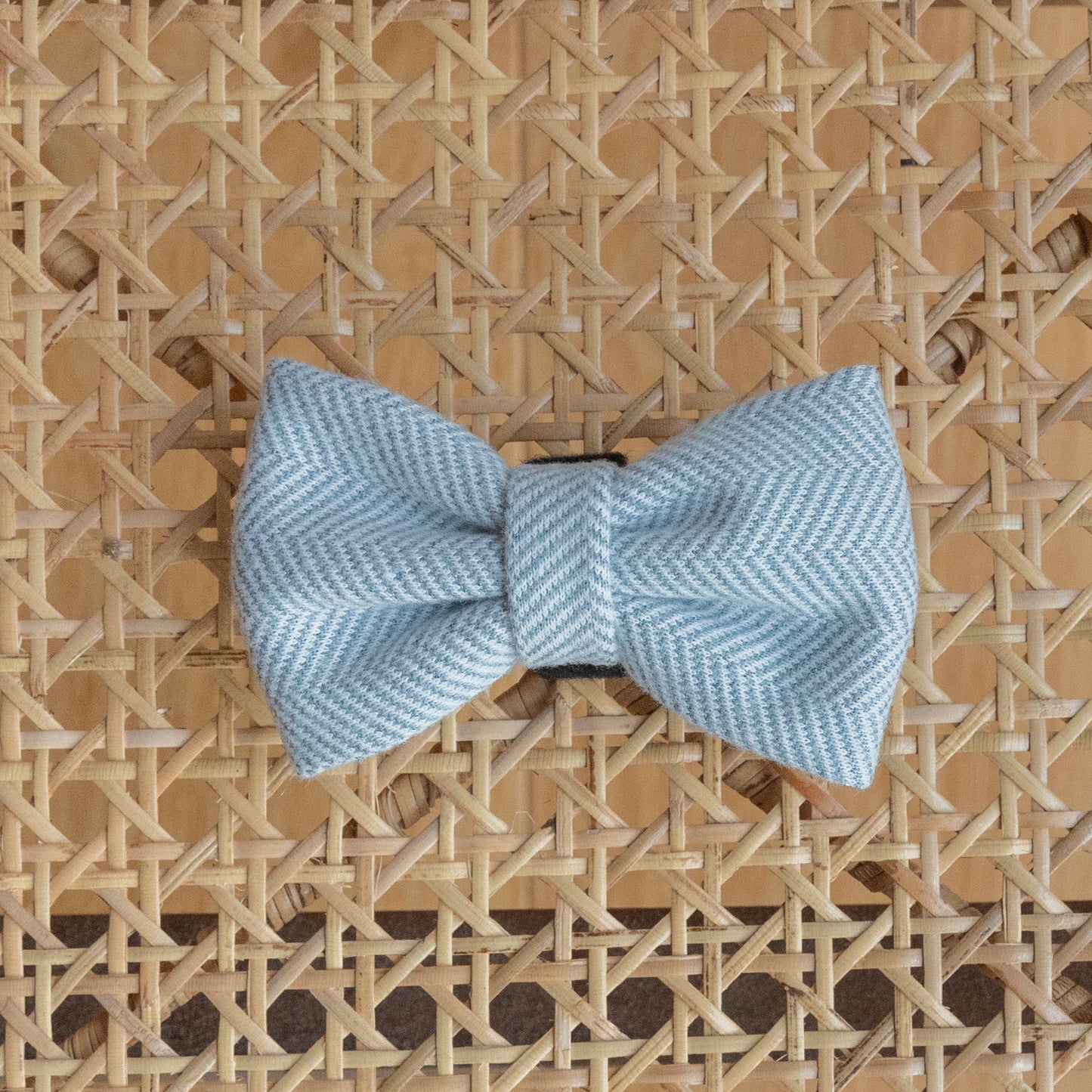 The Sky Tweed Dog Bow Tie, in light blue and crafted from quality materials, rests on a woven lattice-patterned surface. It features a stylish herringbone design that adds a subtle decorative element to the intricate background.