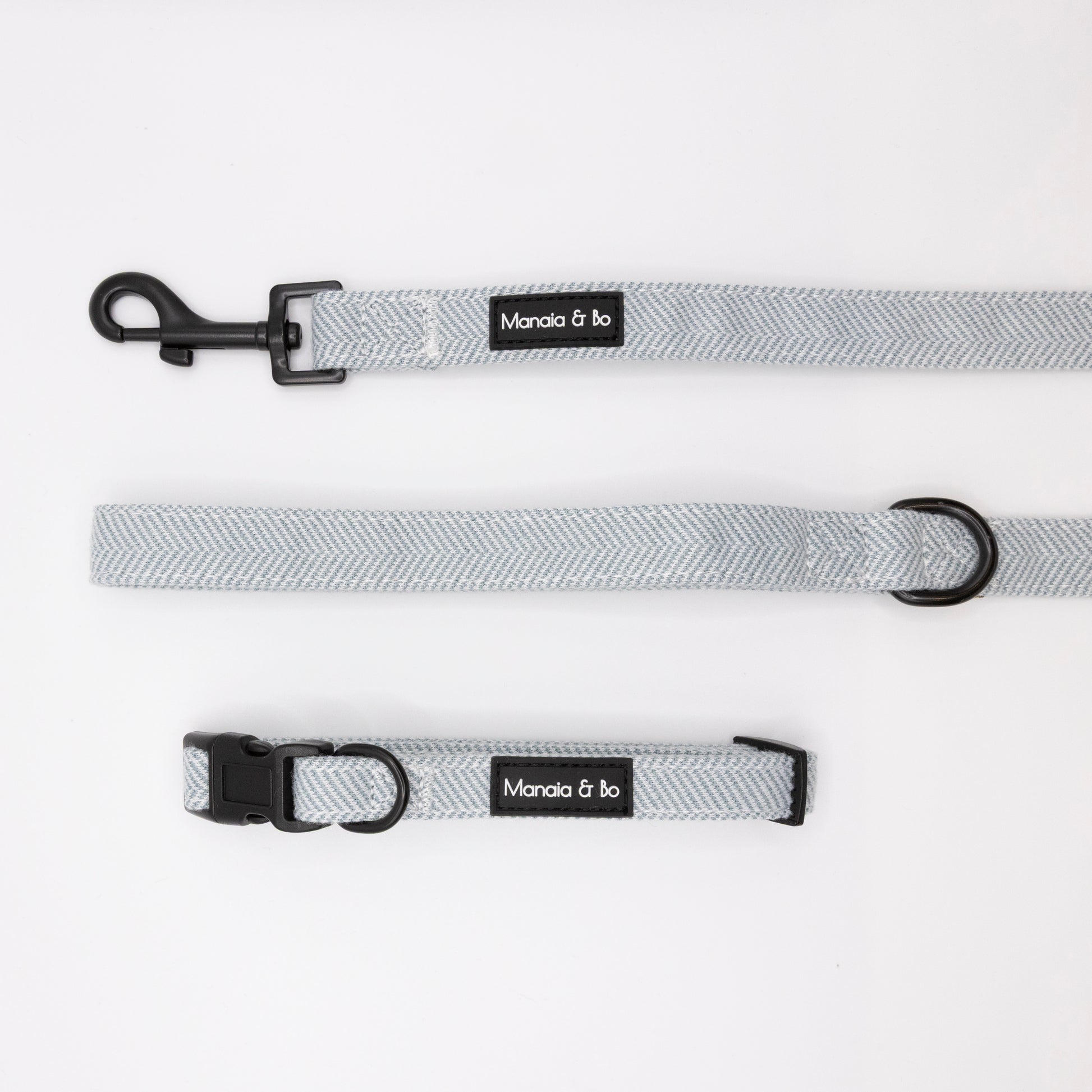 Sky Tweed Collar & Lead set in light gray with black metal hardware is displayed on a white background. Both feature a Mancia & bo label. The leash includes a clasp; the collar has a plastic buckle and metal ring.