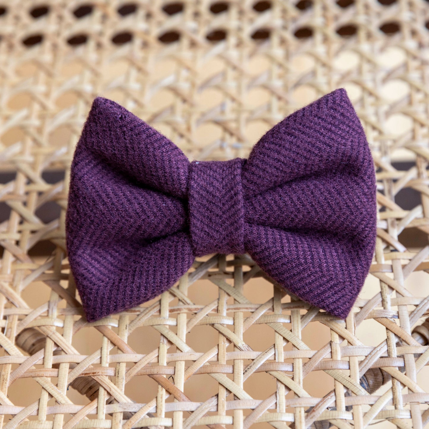 A stylish Wild Plum Tweed Dog Bow Tie, crafted from quality materials, rests on a woven rattan surface, with its textured fabric adding an extra touch of sophistication.