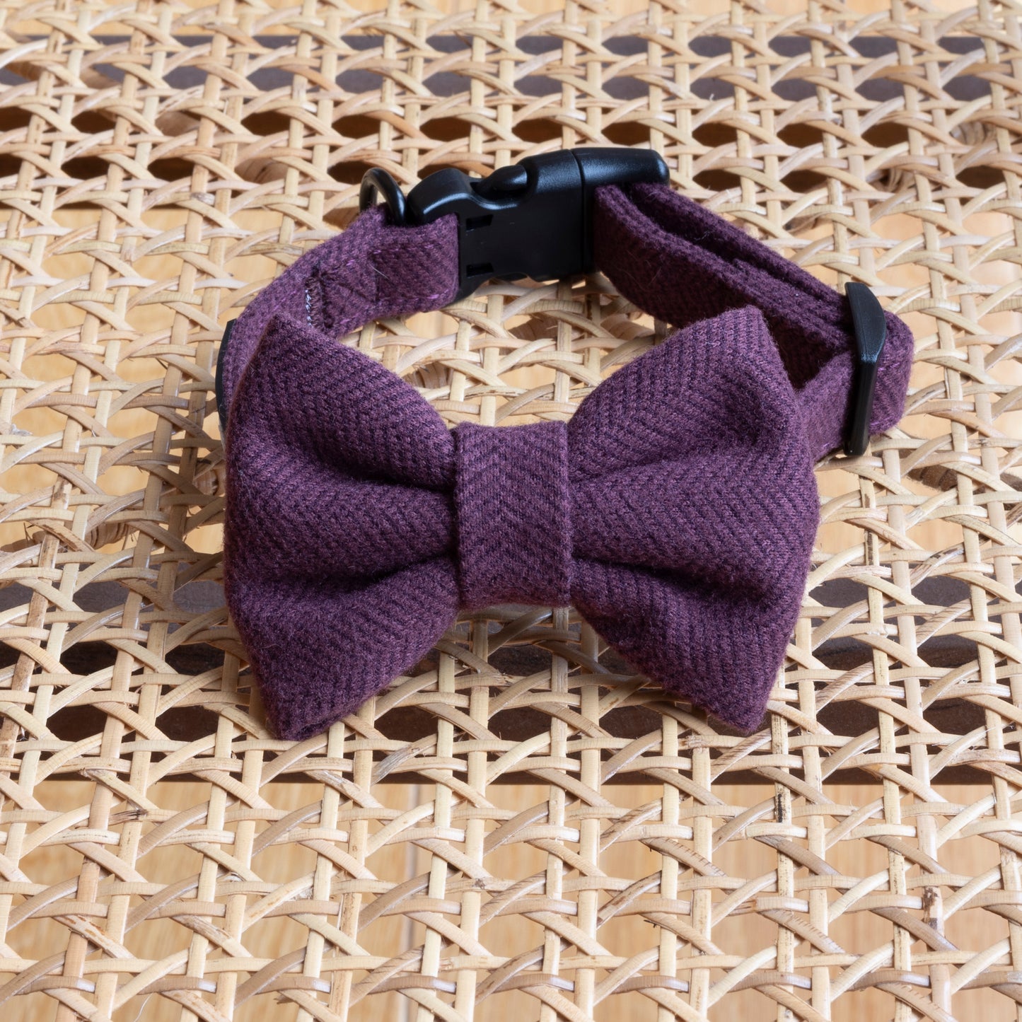 The Wild Plum Tweed Dog Bow Tie, a chic purple fabric accessory crafted from quality materials with a black adjustable strap, elegantly rests on a woven rattan surface attached to a collar.