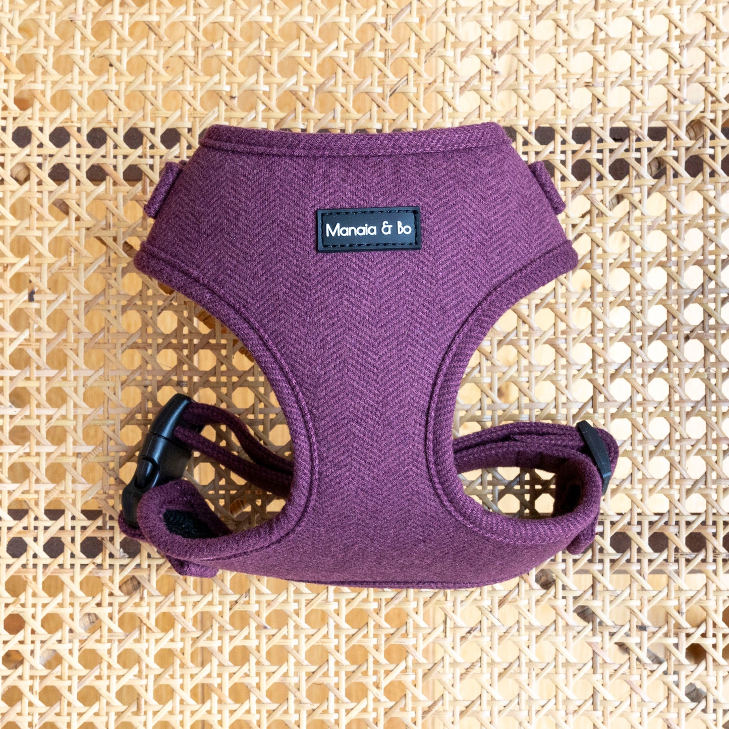 A cozy Wild Plum tweed Dog Harness featuring maroon color, black buckles, and a Manaia & bo tag lies on a woven rattan surface, highlighting its quality materials.