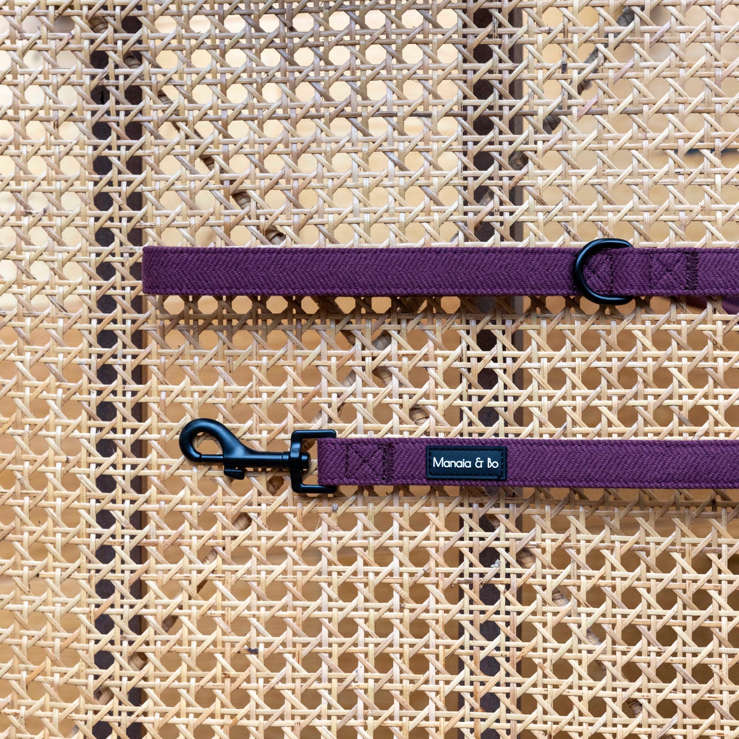 The Wild Plum Tweed Dog Lead, featuring a black clasp and a Monica & Jo label, rests elegantly on the woven rattan surface, highlighting its quality materials.