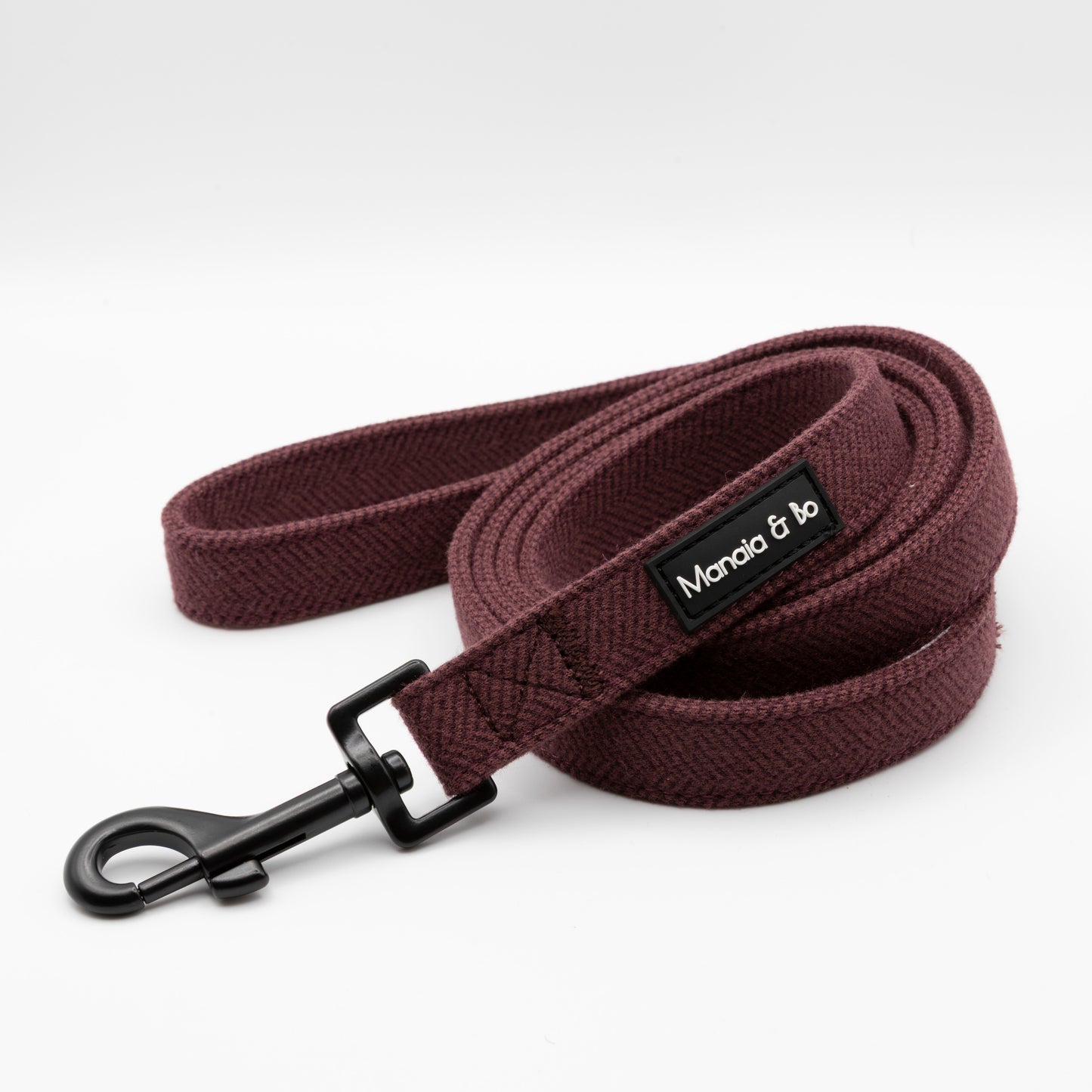 The Wild Plum Tweed Dog Lead features maroon herringbone fabric, a black metal clip, and a black Moncia & Jo label, shown coiled neatly on a white background.