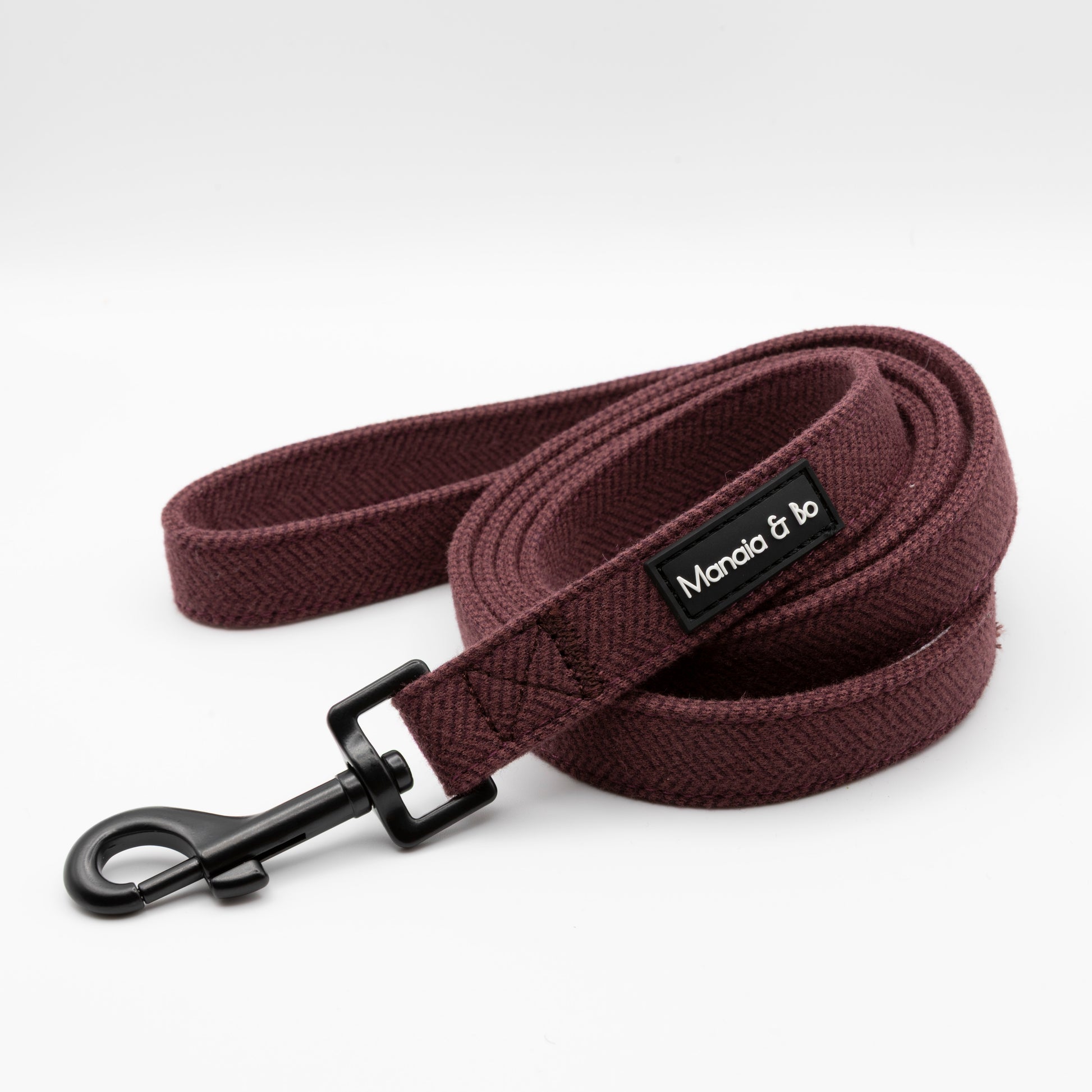The Wild Plum Tweed Dog Lead features maroon herringbone fabric, a black metal clip, and a black Moncia & Jo label, shown coiled neatly on a white background.